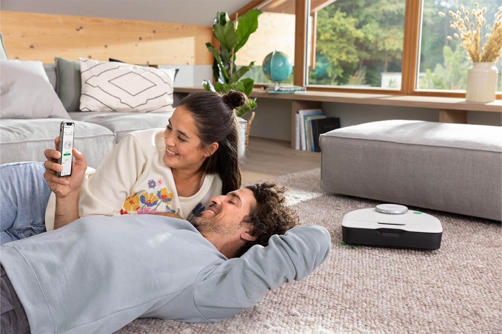Ein fröhliches Paar liegt auf einem Teppichboden im Wohnzimmer und schaut auf die Vorwerk-App. Daneben saugt der neue Vorwerk Saugroboter.