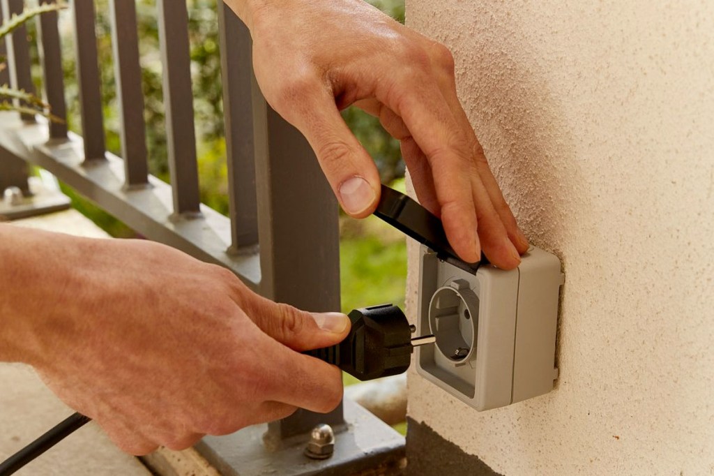 Eine Person steckt einen Schuko-Stecker in eine Außensteckdose.