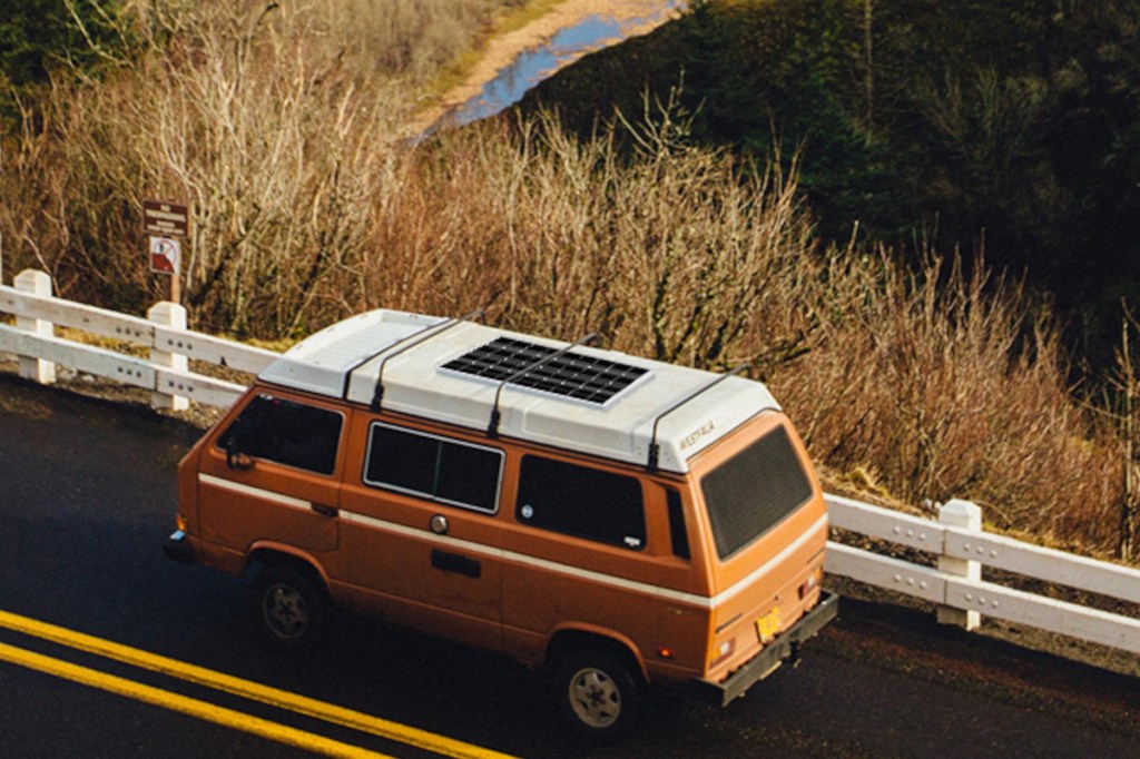 Orangener Camper mit Solarmodul auf Dach auf dunkler Straße umgeben von getrocknetem Gras und Sträuchern