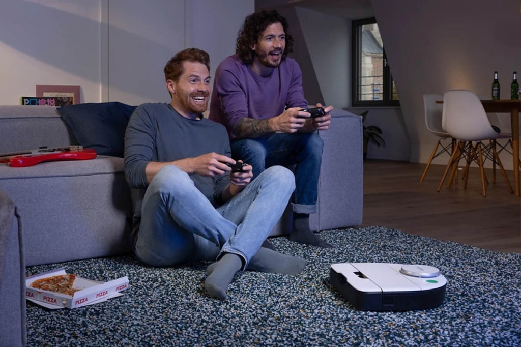Zwei Personen spielen in einem Wohnzimmer auf einer Spielekonsole. Der Kobold VR7 von Vorwerk saugt derweil den Teppich.