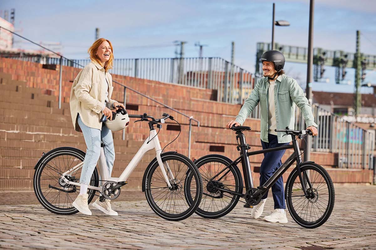 Zwei Menschen stehen neben ihren E-bikes lachend auf einem Platz.