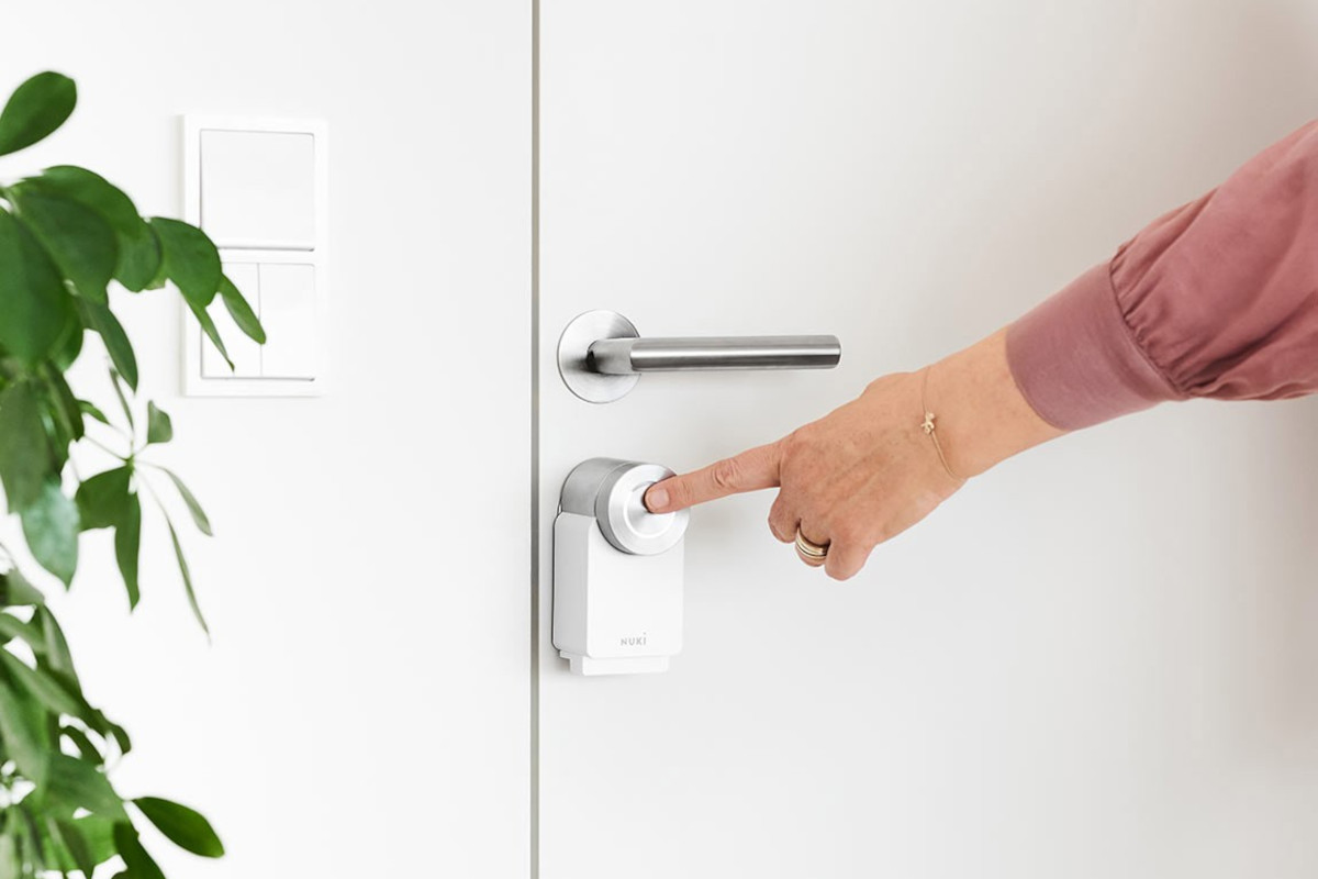 Finger am ausgestreckten Arm berührt weißes Nuki Smart Lock 3.0 Pro an weißer Tür neben weißer Wand und links grüner Pflanze