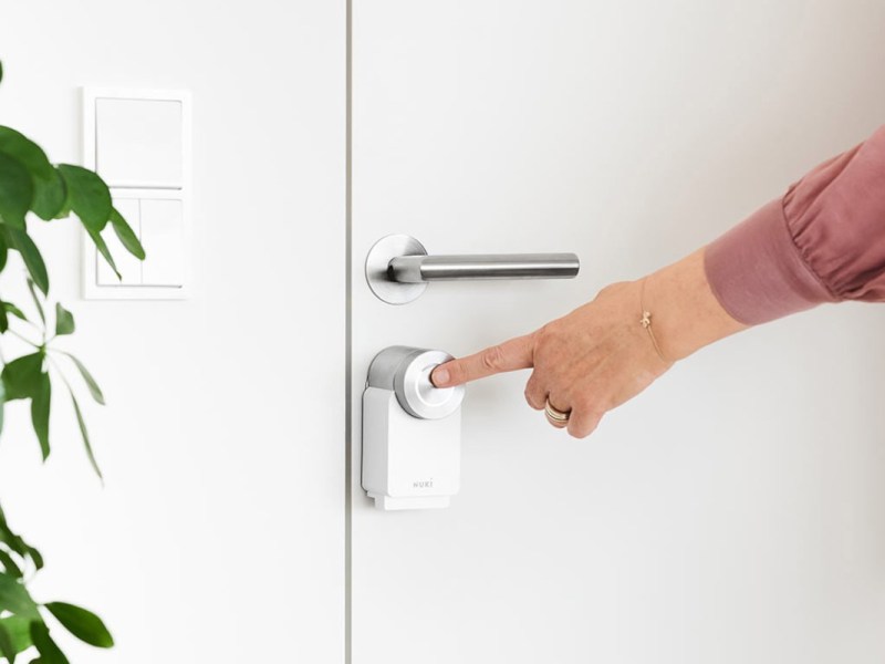 Finger am ausgestreckten Arm berührt weißes Nuki Smart Lock 3.0 Pro an weißer Tür neben weißer Wand und links grüner Pflanze