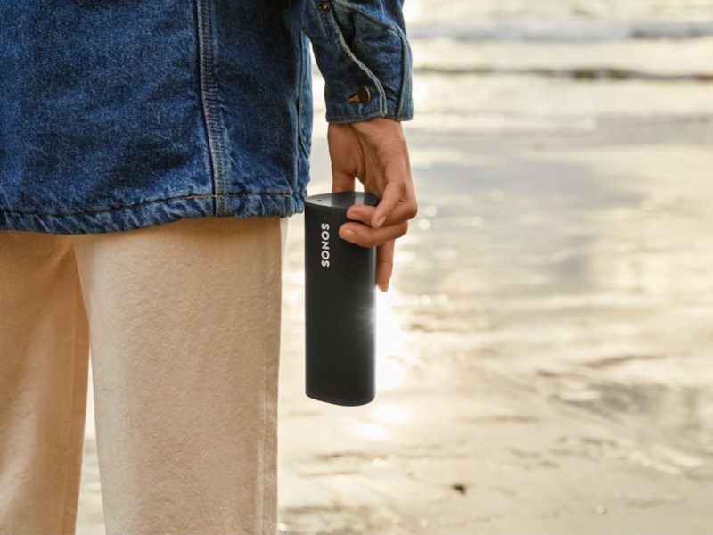 Person in beiger Hose und Jeansjacke hält schwarzen Sonos-Speaker in der Hand und steht am Strand