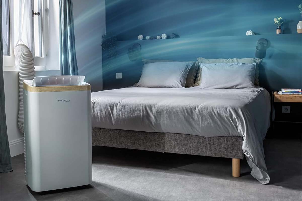 Bett im Hintergrund, im Vordergrund steht ein mobiles Klimagerät, dass mit Hilfe einer Grafik optisch sichtbar macht, dass es Luft versprüht