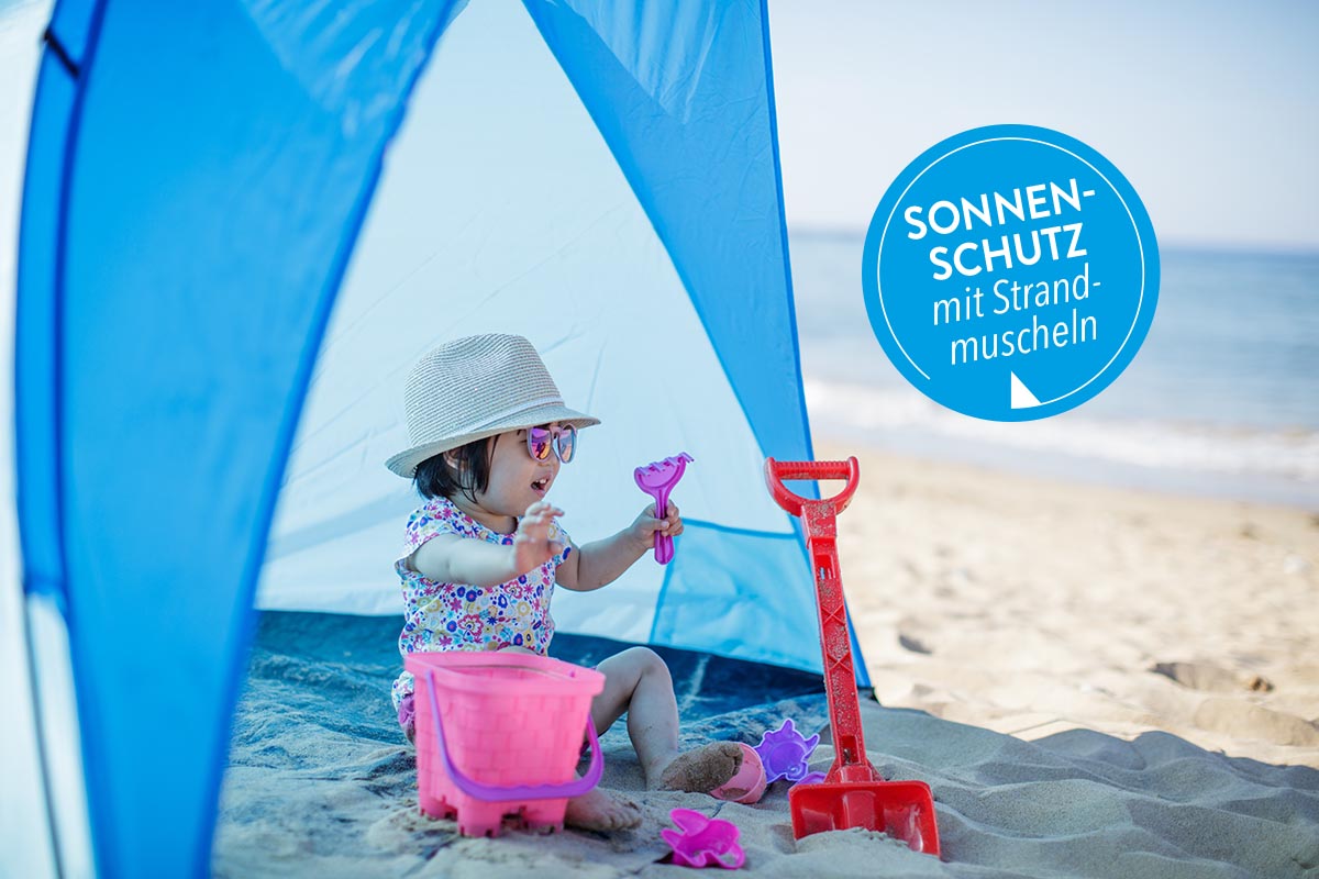 Ein Kind sitzt am Strand in einer Strandmuschel.