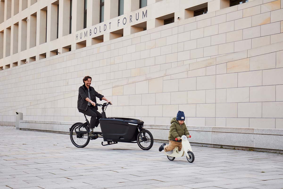 Mann fährt mit einem Cargo-E-Bike über einen Platz, ein kleines Kind fährt vor ihm mit einem Laufrad