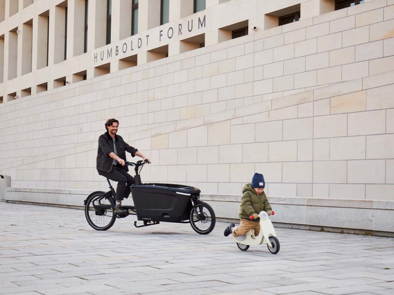 Mann fährt mit einem Cargo-E-Bike über einen Platz, ein kleines Kind fährt vor ihm mit einem Laufrad