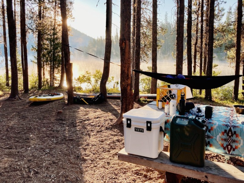 Waldlichtung bei Sonnenschein mit aufgebauten Campingtisch und Bänken auf dem Boxen und Trinkflaschen stehen, im Hintergrund eine Hängematte