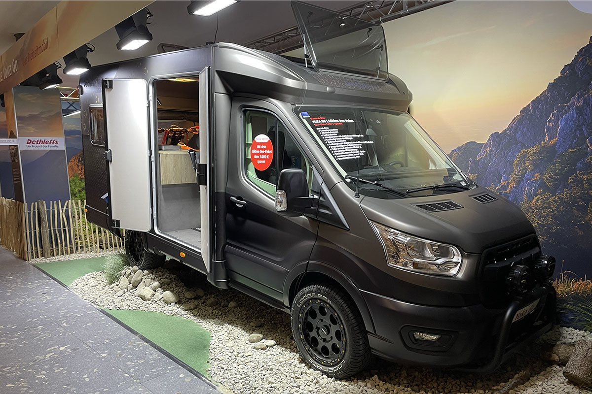 Camper Van mit Outdoor-Eigenschaften wie hohem Radstand und Crossreifen auf Messestand. Seitlich von vorn abgelichtet mit geöffneter Tür in den Wohnraum.