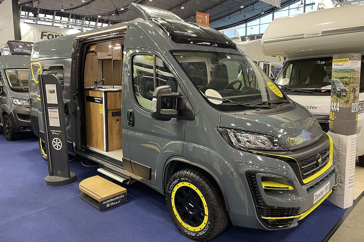 Camper Van mit Outdoor-Eigenschaften wie hohem Radstand und Crossreifen auf Messestand. Seitlich von vorn abgelichtet mit geöffneter Tür in den Wohnraum.