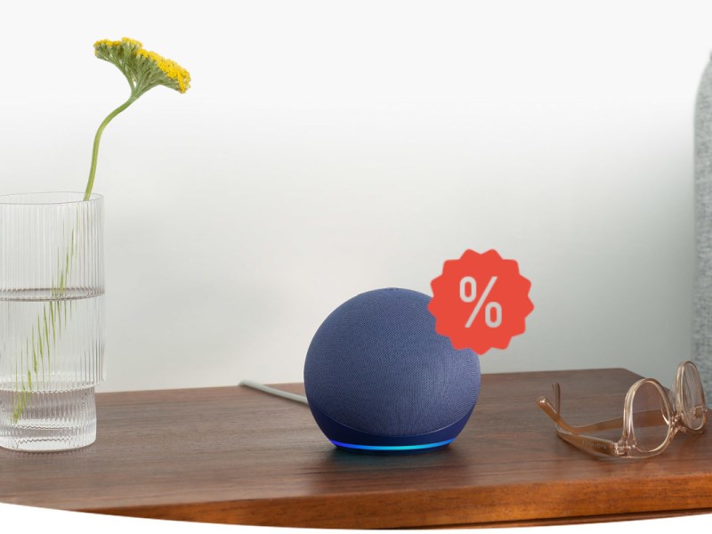 Dunkelblauer Echo Dot 5 kugelförmiger Lautsprecher auf Holzbeistelltisch neben Glas mit gelber Blume und brauner Brille vor weißer Wand