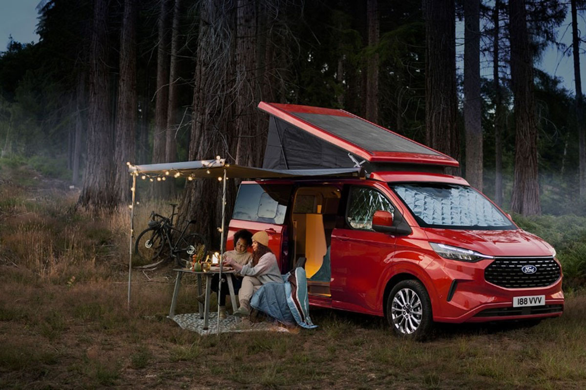 Camper Van-Alternative zum VW California: Ein Ford Nugget steht mit aufgestelltem Hochdach vor einem Waldstück. Davor die beiden Campenden beim Essen.