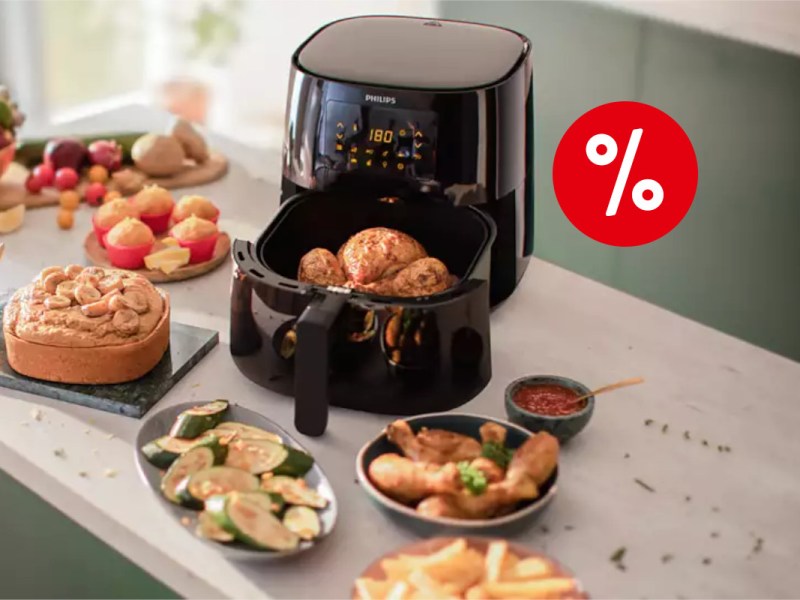 Schwarze Philips Essential Airfryer XL Heißluftfritteuse geöffnet mit gegartem Hähnchen drin von oben auf heller Küchenarbeitsfläche zwischen Tellern und Schüsseln mit anderen frittierten Lebensmitteln, oben rechts ein rotes Prozentzeichen