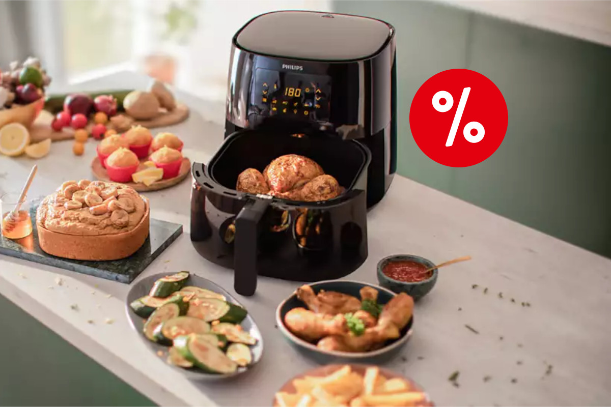 Schwarze Philips Essential Airfryer XL Heißluftfritteuse geöffnet mit gegartem Hähnchen drin von oben auf heller Küchenarbeitsfläche zwischen Tellern und Schüsseln mit anderen frittierten Lebensmitteln, oben rechts ein rotes Prozentzeichen