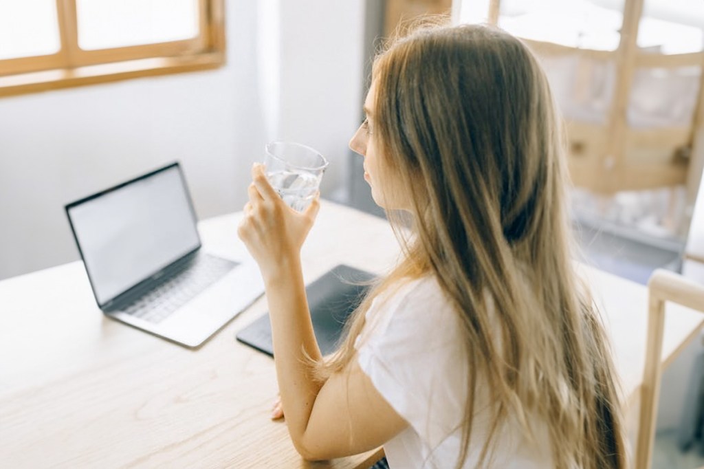 Una mujer se sienta en un escritorio con una computadora portátil y bebe agua de una taza.