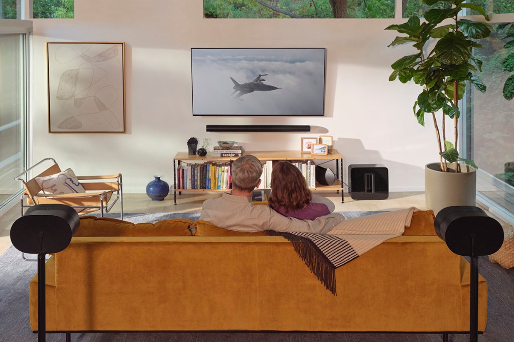 Frau und Mann sitzen auf einem Sofa vor einem Flachbild-TV. Hinter ihnen befinden sich rechts und links zwei Rücklautsprecher für die Sound-Wiedergabe beim TV-schauen.