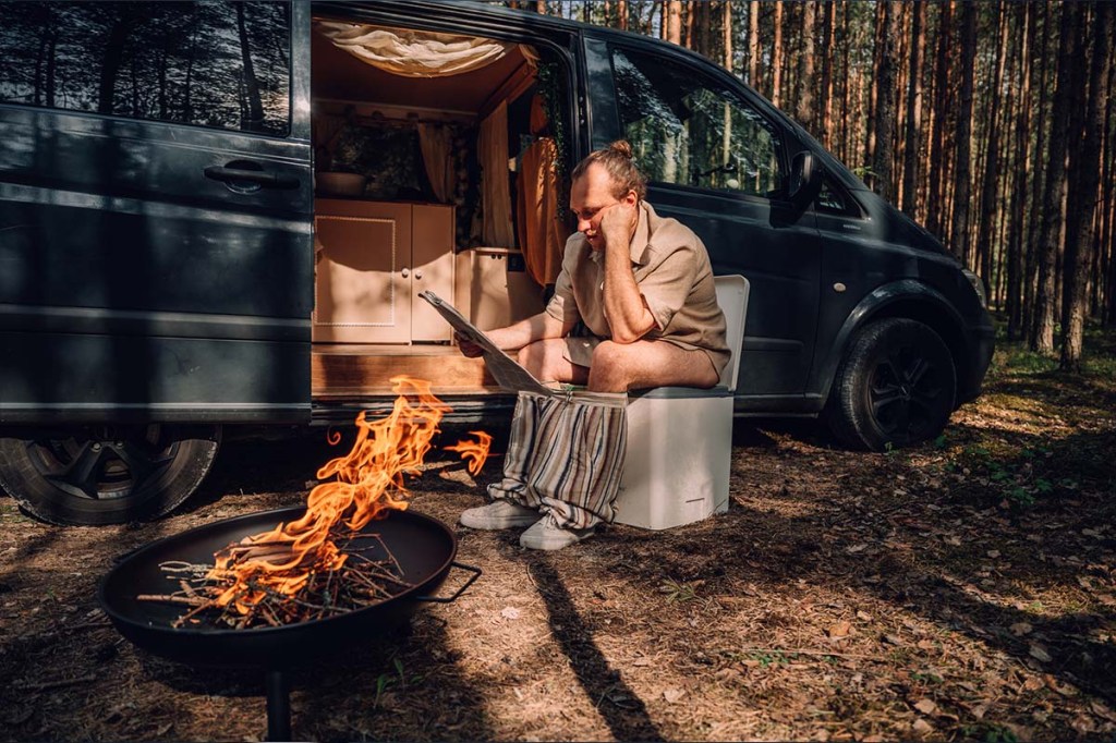 Mann sieht im Wald auf seiner Trenntoilette, hinter ihm sein Camper, neben ihm eine Feuerschale