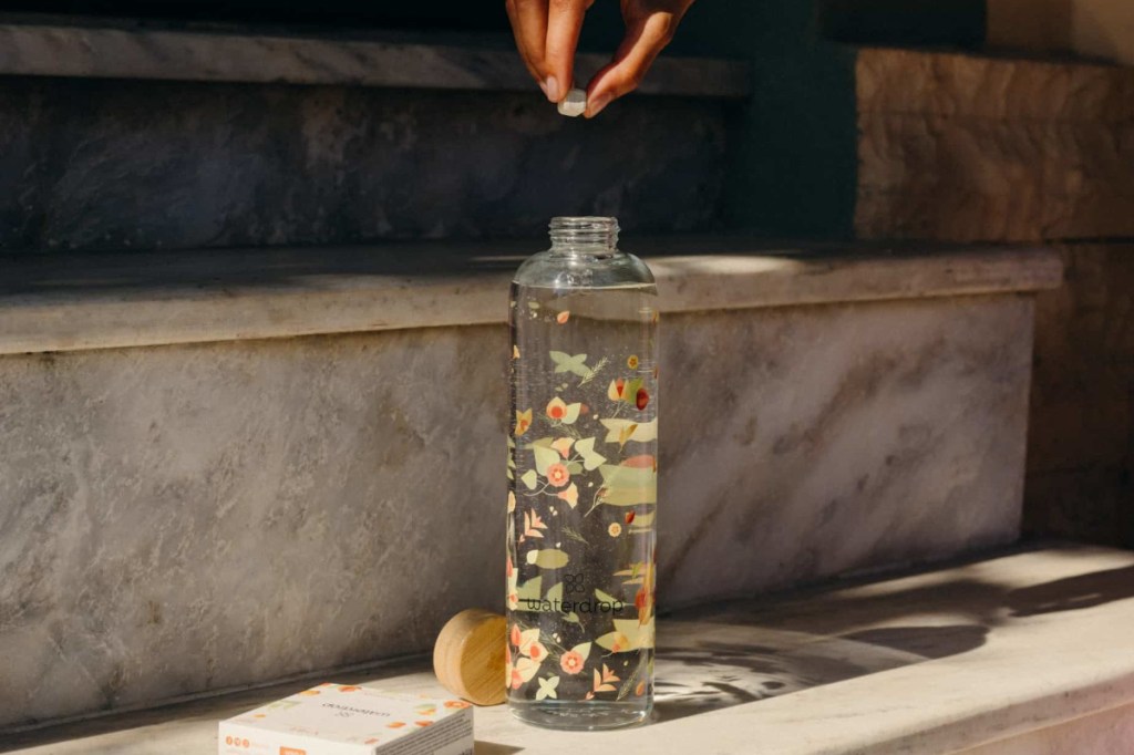 Waterdrop-Trinkflasche durchsichtig mit buntem Blumenmuster auf heller Steintreppe dadrüber Finger die Tablette halten