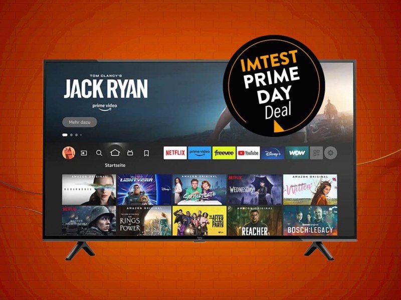 Amazon Fire TV Fernseher von vorne mit geöffneter Mediathek auf orangenem Hintergrund mit schwarzem Button "IMTEST Prime Day Deal"