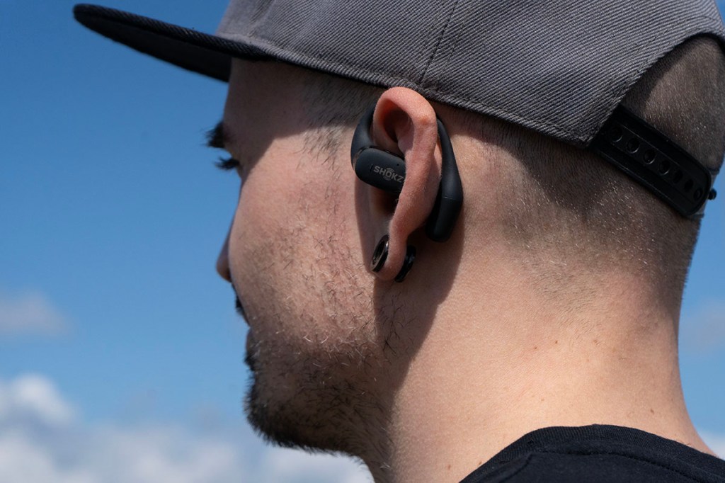 Eine Person trägt den Ohrhörer Shokz OpenFit, von schräg hinten fotografiert, davor blauer Himmel.