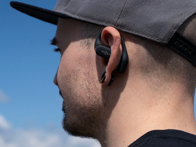Eine Person trägt den Ohrhörer Shokz OpenFit, von schräg hinten fotografiert, davor blauer Himmel.