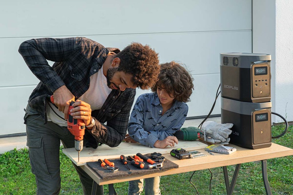 Ein Mann und ein Junge arbeiten im Garten mit einer Bohrmaschine. Auf dem Tisch steht daneben eine Powerstation mit Zusatz-Akku.