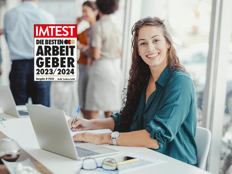 Lächelnde Frau an weißem Schreibtisch und aufgeklapptem weißen Notebook, im Hintergrund stehende Leute, Links oben IMTEST Siegel "Die besten Arbeitgeber 2023/2024"