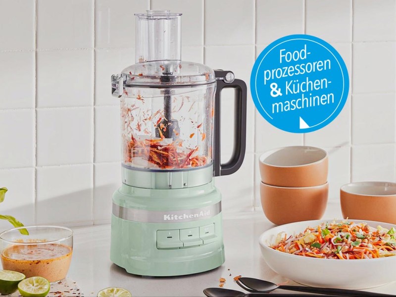 Mintgrüner zylinderförmiger Food Processor mit durchsichtigem Aufsatz in dem rotes Lebensmittel zerhackt ist auf weißem Tisch neben Teller und Schüsseln vor weißer Wand mit blauem Button rechts oben "Food-Prozessoren und Küchenmaschinen"