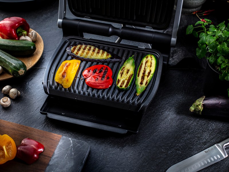 Aufgeklappter rechteckiger schwarzer Tefal Optigrill mit gerilltem Gemüse drauf auf dunkler Arbeitsfläche mit Brett, Messer und Gemüseresten