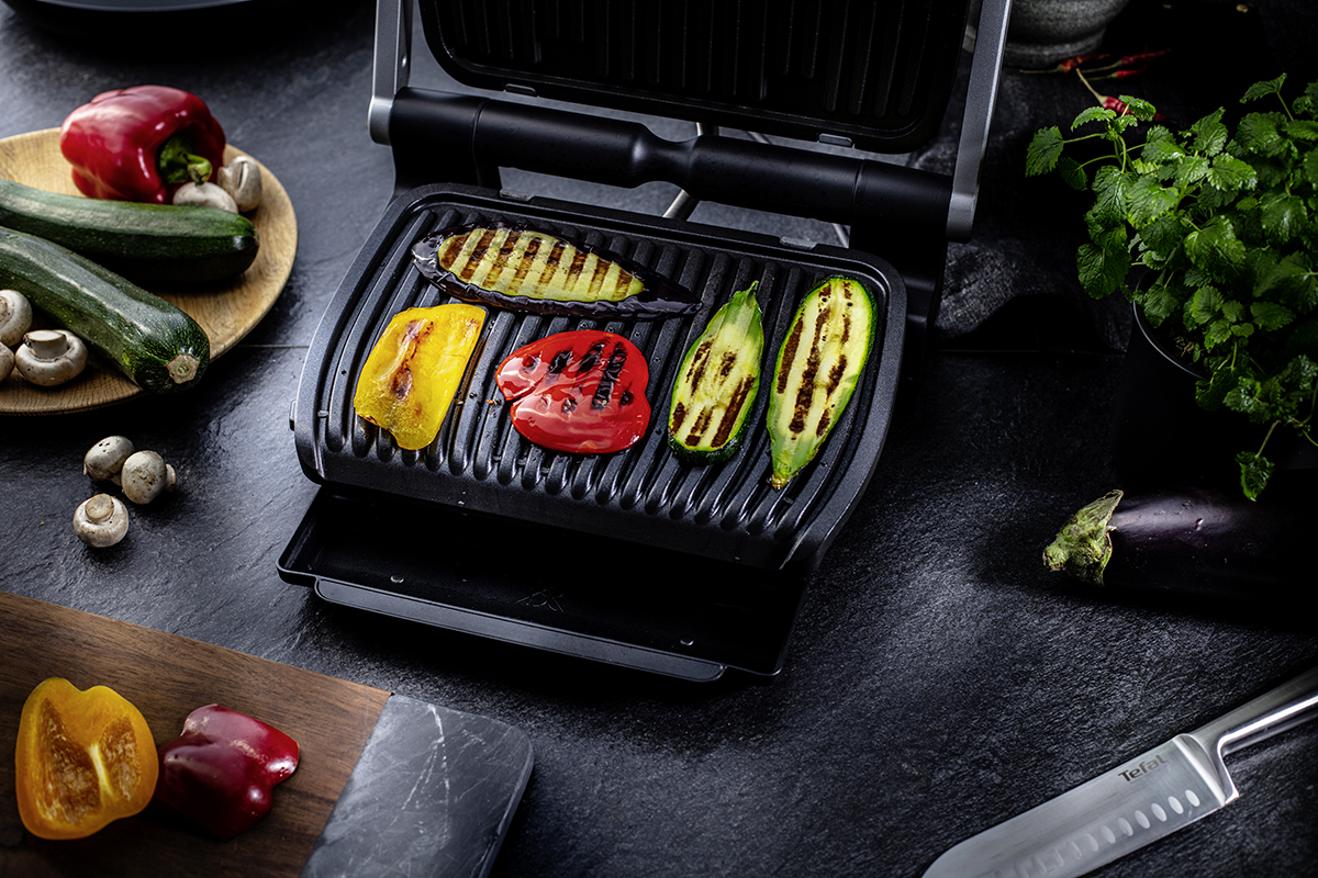 Aufgeklappter rechteckiger schwarzer Tefal Optigrill mit gerilltem Gemüse drauf auf dunkler Arbeitsfläche mit Brett, Messer und Gemüseresten