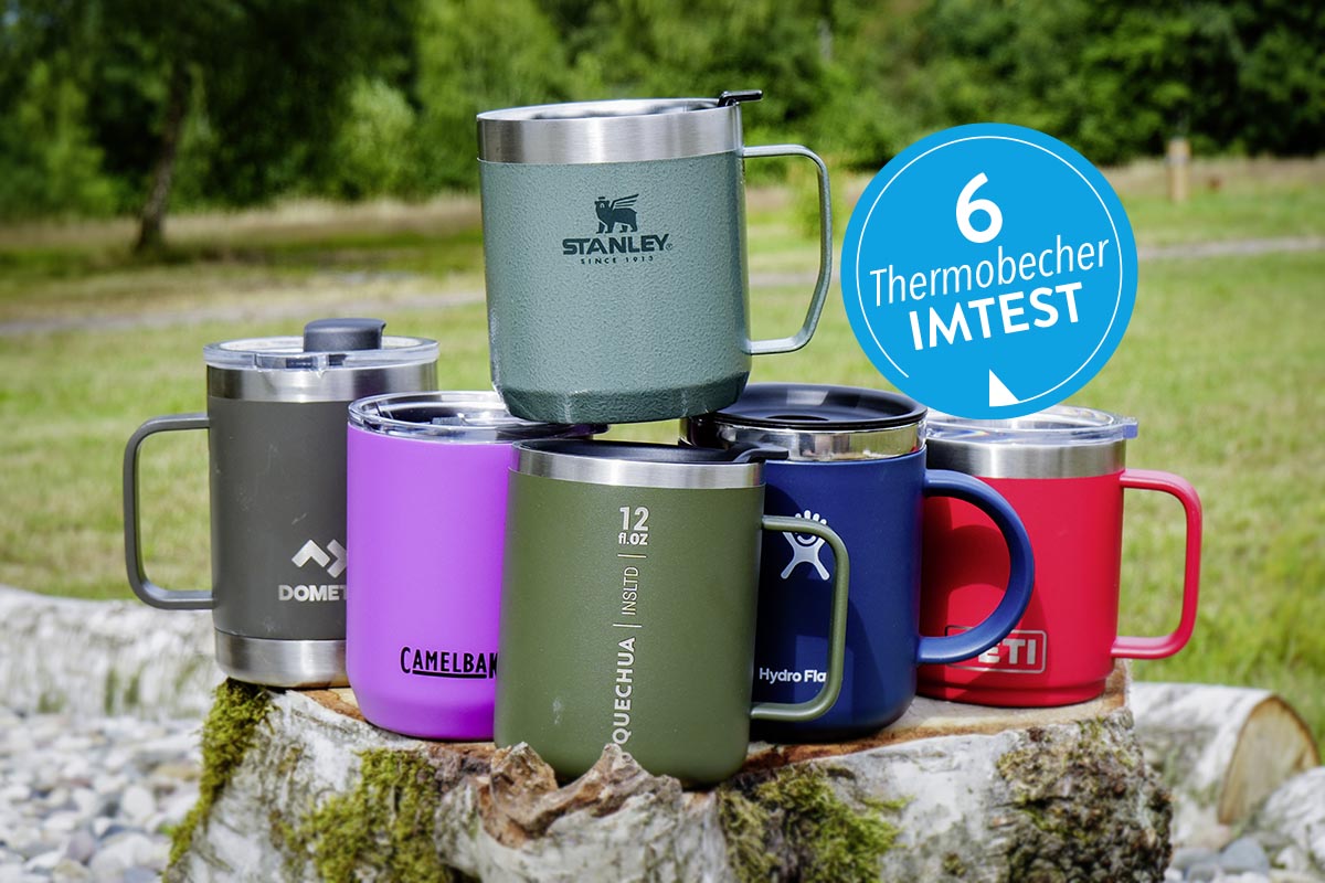 Sechs Thermobecher in Tassenform in unterschiedlichen Farben aufgereiht, einer auf die anderen gestellt auf einem Felsen vor grüner Wiese