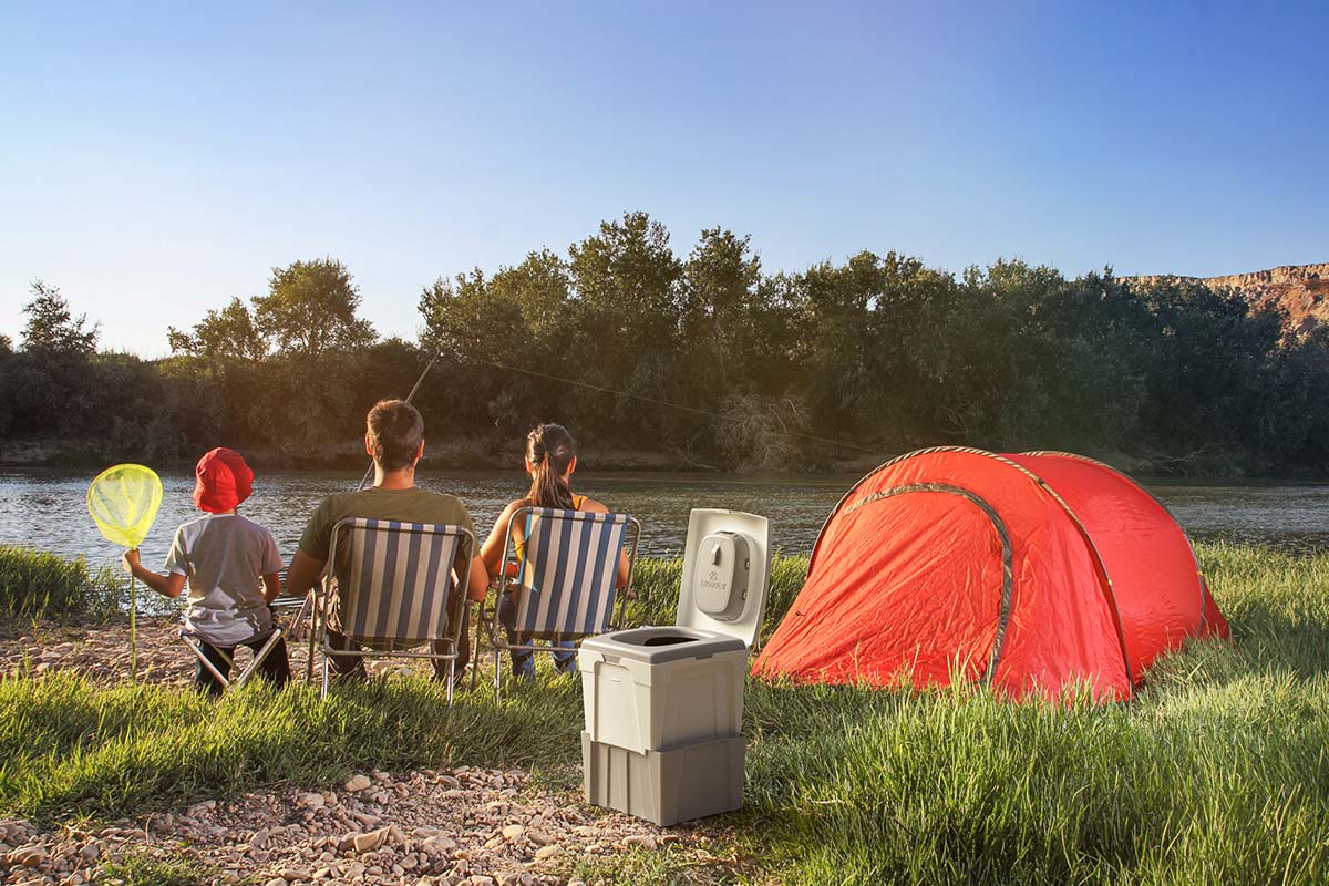 Drei Personen von hinten auf Stühlen an einem SEe sitzend, neben ihnen ihr Zeit, dahinter eine Trenntoilette