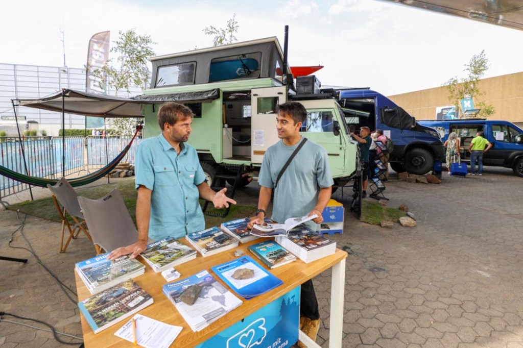 Zwei Personen unterhalten sich an einem Tisch mit Info-Material. Im Hintergrund sind Campervans.