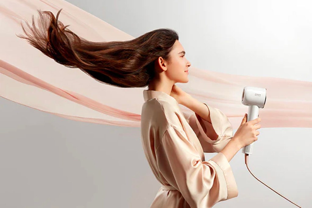 Eine Frau föhnt sich die Haare mit dem Dreame Hair Glory. Im Hintergrund weht ein rosa-farbenes Tuch.