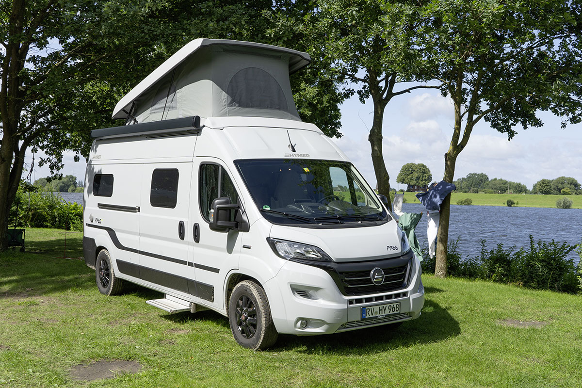 Camping-Kastenwagen Hymer Free 600 mit aufgestelltrem Hochdach auf Campingplatz – im Hintergrund fließt ein Fluss.