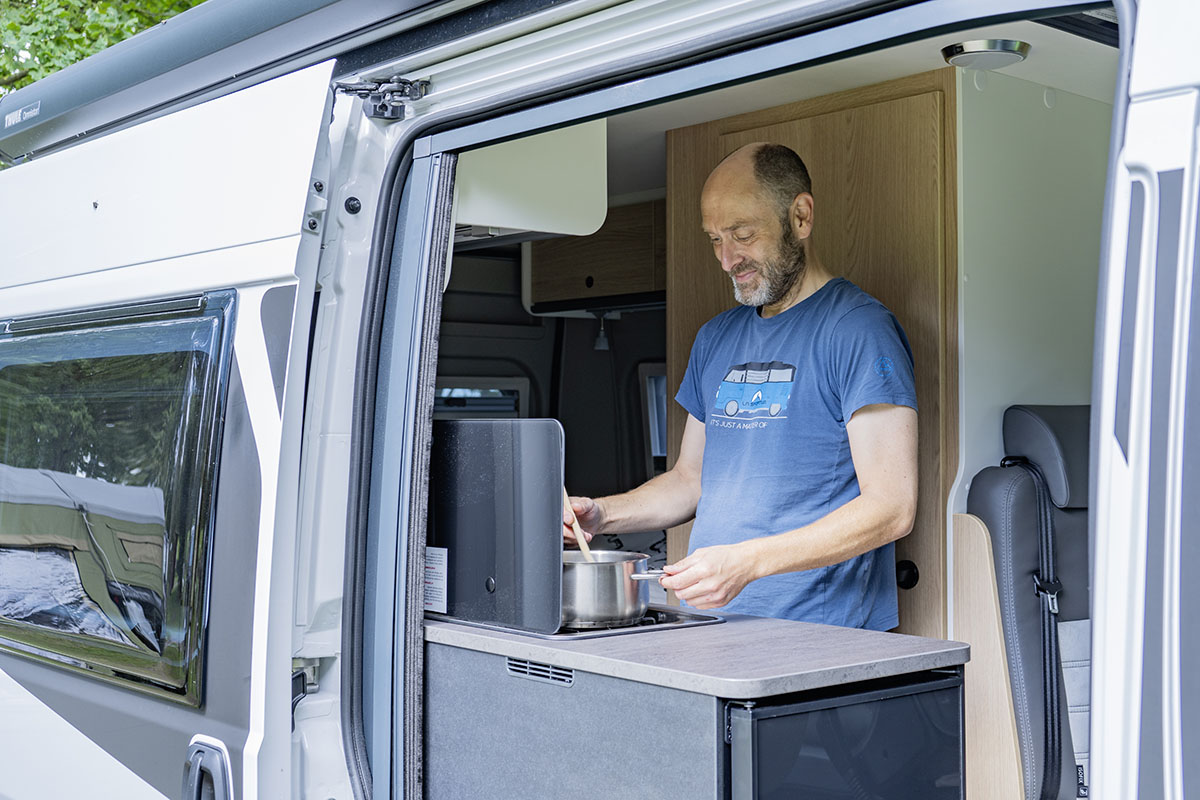 Mann kocht bei geöffneter Seitenschiebetür im Camping-Kastenwagen Sunlight Cliff 540 RT Adventeure Edition.
