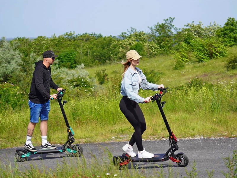 Zwei Personen fahren auf einer Straße im Grünen mit E-Scootern von IO Hawk.