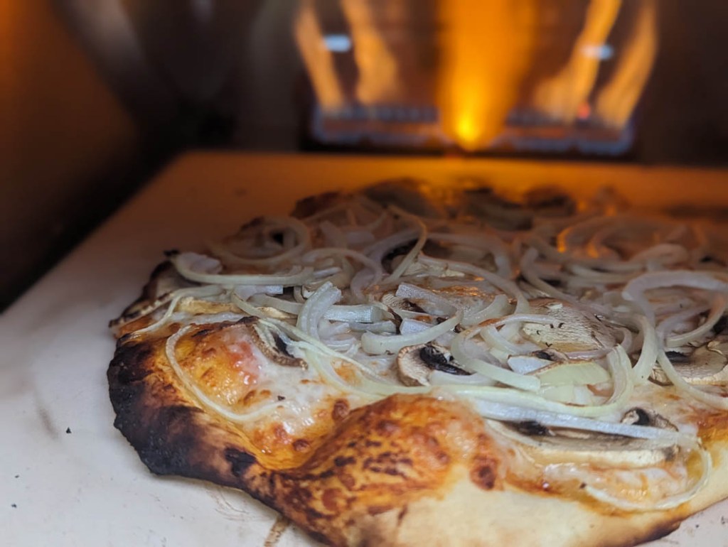 Ein Pizza im Nero, auf der linken Seite sehr dunkel bis verbrannt. Im Hintergrund sind die Flammen des Brenners zu erkennen. 