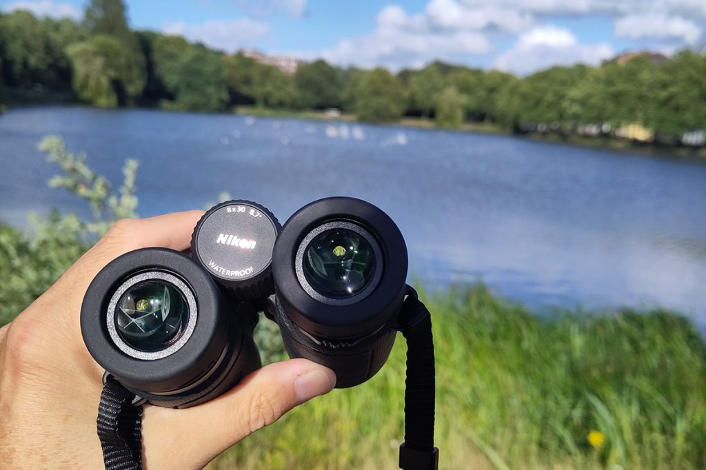 Bild von einem Nikon-Fernglas in der Hand, im Hintergrund ist ein See zu sehen.