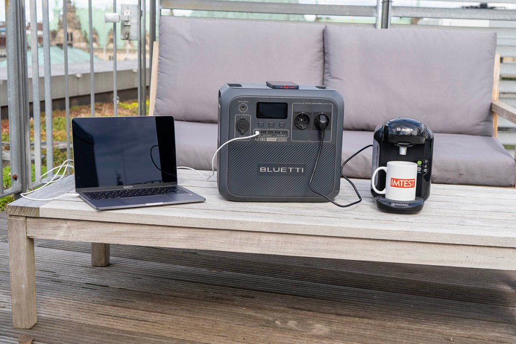 Die Bluetti AC180 mit mehreren Endgeräte (Laptop, Smartphone, Kaffeemaschine) auf einer Dachterrasse mit Sitzmöbeln.