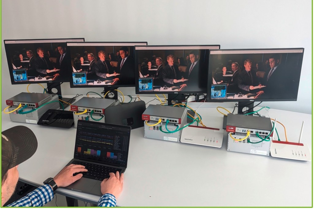Vier kleine Computer-Bildschirme in Reihe aufgestellt zeigen gleiches Bild von Series davor Boxen und Router und davor Mann mit Cappie von hinten am aufgeklapptem Notebook