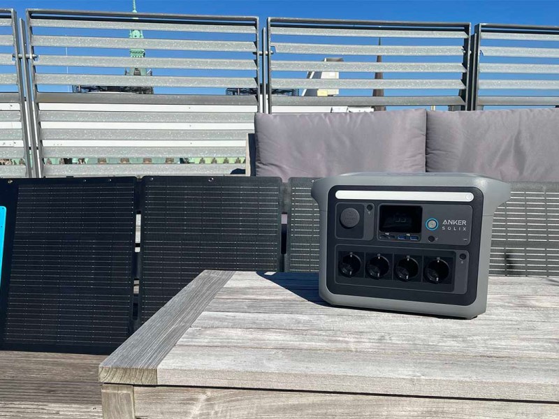 Die Anker Solix C1000 auf einem Tisch in der Sonne. Im Hintergrund steht ein Solarpanel und ein Sitzmöbel.