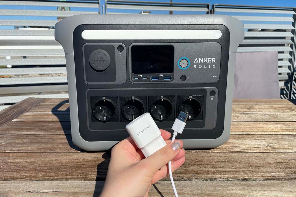 Eine Hand hält ein USB-Kabel und ein passendes Netzteil vor eine Anker-Powerstation.