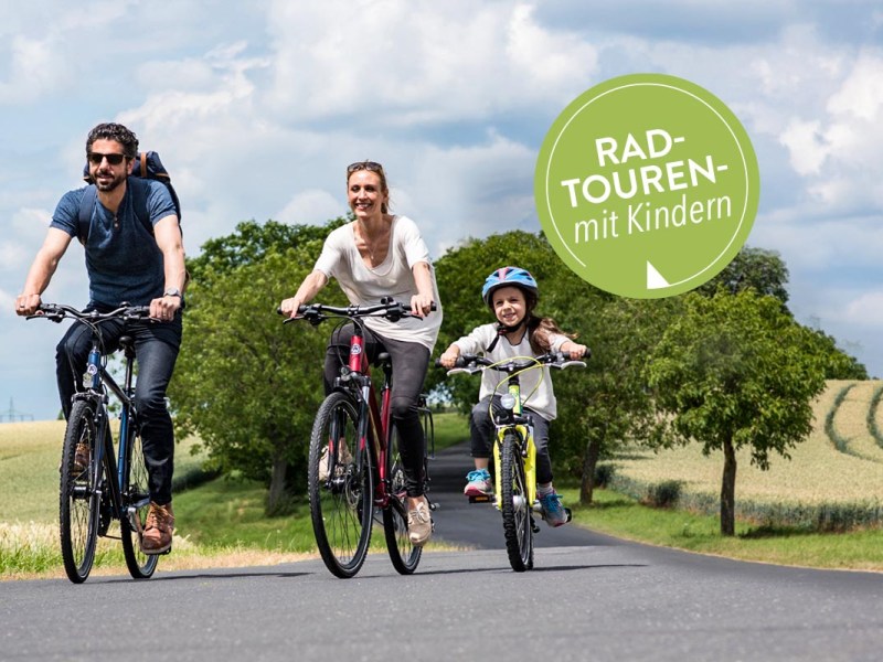 Eine dreiköpfige Familie fährt mit dem Fahrrad eine Straße im Grünen entlang