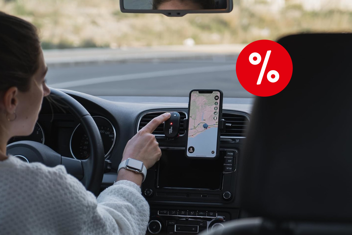 Frau am Lenkrad im Auto in weißem Pulli mit dunklen Haaren streckt Finger nach kleinem schwarzem Gerät an Lüftung aus, daneben ein Smartphone mit Navigation, ein rotes Prozentzeichen rechts oben