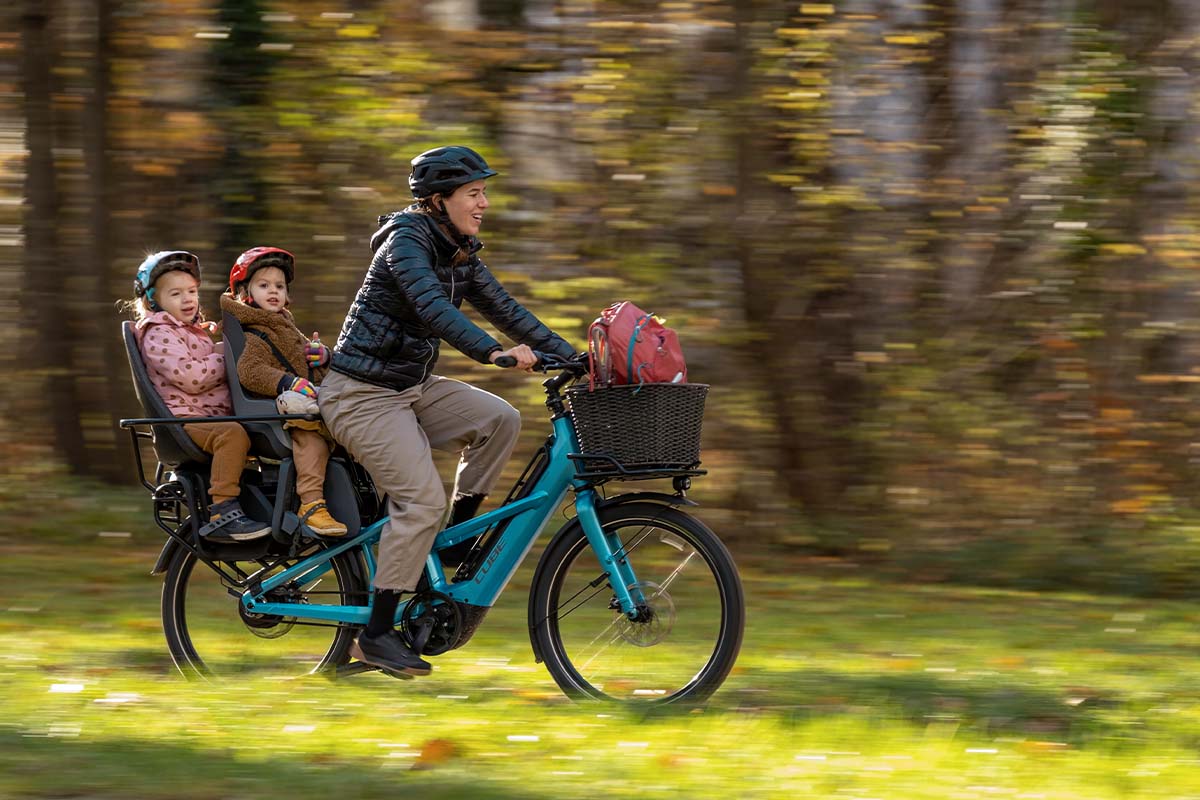 Frau fährt mit einem Fahrrad durch einen Park, auf ihrem Gepäckträger sitzen zwei Kinder in einem Kindersitz
