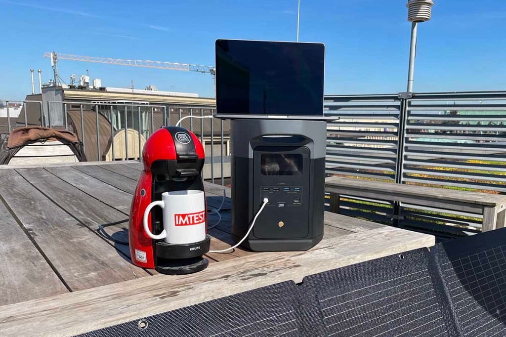 Die Delta 2 Max auf einem Tisch in der Sonne. Darauf ein Laptop und daneben eine Kaffeemaschine mit IMTEST-Tasse.