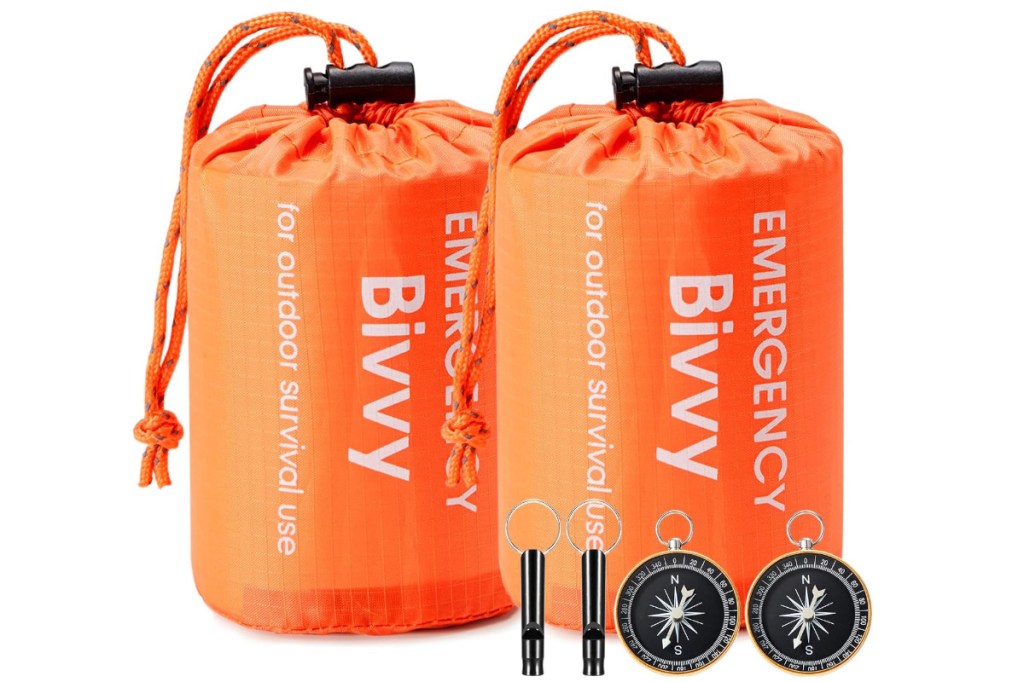 Zwei orangene eingepackte Biwacksäcke nebeneinander, davor zwei Signalpfeifen und dunkle Kompasse auf weißem Hintergrund