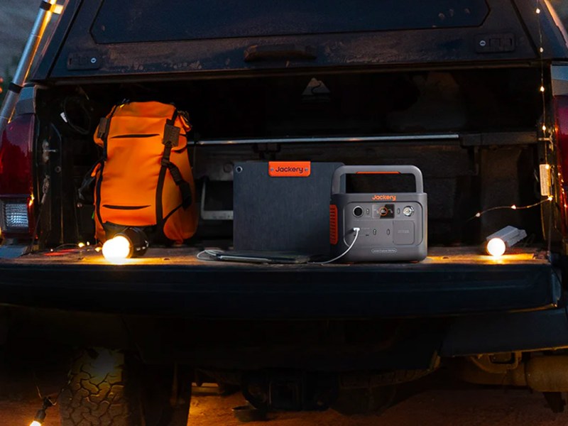 Die Explorer 300 Plus in einem geöffneten Kofferraum neben dem passenden Solarpanel, einem Rucksack und mit einer Lichterkette beleuchtet.